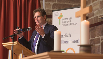 Pastor Thorsten Graff, Leiter des Konzernbereichs Seelsorge-Theologie-Ethik, spricht am Rednerpult auf der Bühne mit Kerzen und einem Holzkreuz beim Zusammenschluss von Diakonie Werratal und Immanuel Diakonie Südthüringen im Kressehof im Meininger Ortsteil Walldorf 